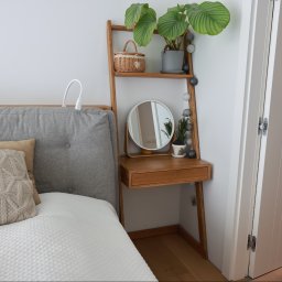 Wojciech Raś Twój Wymiar Pracownia Mebli - Drewniana drabina-regał z lustrem i szufladą, ustawiona przy łóżku z szarym zagłówkiem. Na półkach koszyk, roślina w doniczce i dekoracje. Białe ściany i drzwi.