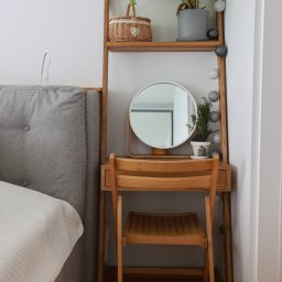 Wojciech Raś Twój Wymiar Pracownia Mebli - Drewniana toaletka z okrągłym lustrem i krzesłem w jasnym wnętrzu. Na półkach dekoracje: roślina w doniczce, koszyk i girlanda świetlna. Obok wezgłowie łóżka.