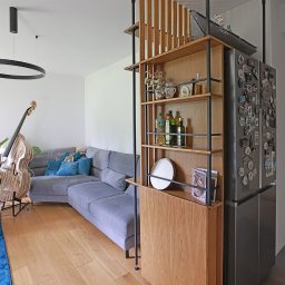 Wojciech Raś Twój Wymiar Pracownia Mebli - Nowoczesny salon z drewnianą zabudową regałową, dzielącą przestrzeń. Widoczny szary narożnik, kontrabas i okrągła lampa. Parkiet z naturalnego drewna.