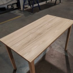 Wojciech Raś Twój Wymiar Pracownia Mebli - Drewniany stół z jasnego drewna w warsztacie w Gdańsku. Minimalistyczny design, widoczne słoje drewna. Stół na czterech nogach, surowy beton na podłodze.