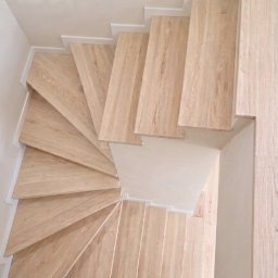 Twój Wymiar Pracownia Mebli - Spiralne schody drewniane z jasnego drewna, widok z góry, minimalistyczny design z białymi listwami przypodłogowymi, oświetlenie naturalne.