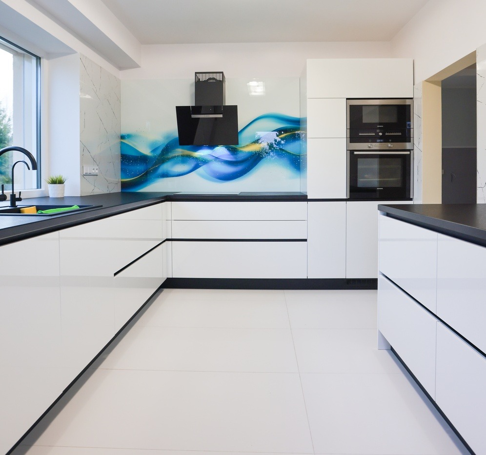 Nowoczesna, biała kuchnia z czarnymi blatami i abstrakcyjnym panelem szklanym. Minimalistyczny design, jasne płytki podłogowe, wbudowane sprzęty AGD.