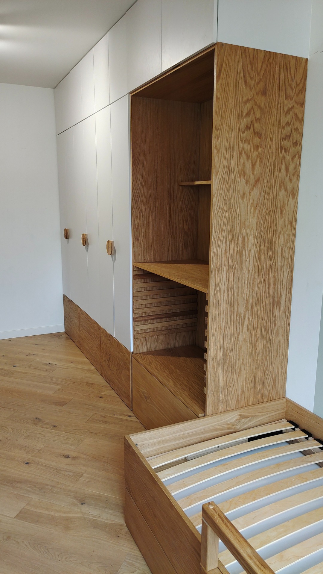 Biało-drewniana szafa wnękowa z otwartą półką i drewnianym łóżkiem bez materaca, na parkiecie. Widoczne detale wykończenia i naturalna faktura drewna.