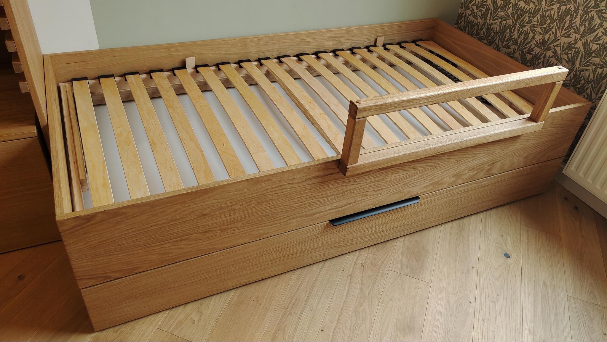 Drewniane łóżko z szufladami i barierką ochronną, wykonane na wymiar. Widoczny stelaż z listew i naturalny odcień drewna dębowego. Minimalistyczny design.