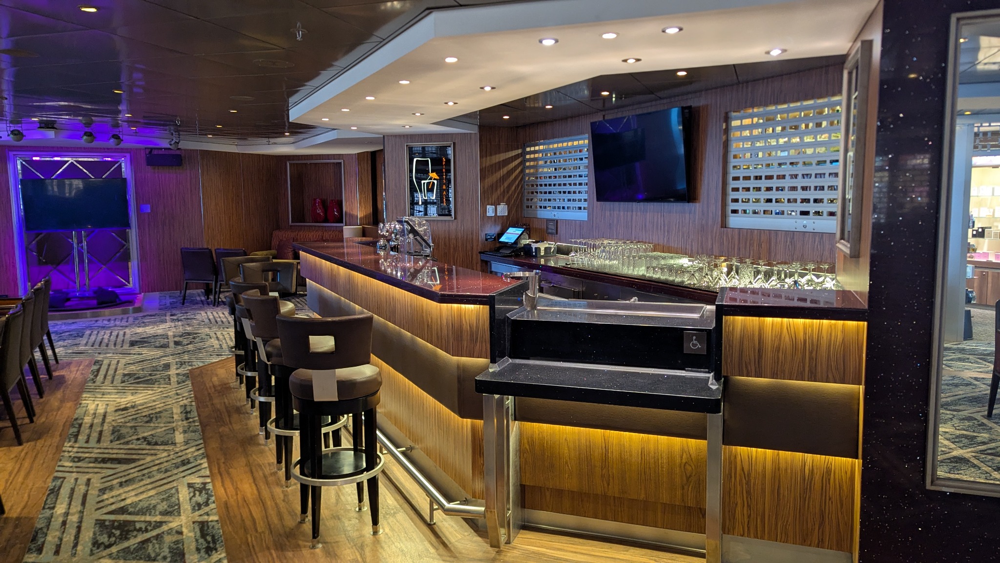 Nowoczesny bar z podświetlanym frontem, ciemnym blatem i rzędem hokerów. W tle scena z fioletowym oświetleniem i ściana z ekspozycją. Eleganckie wykończenie wnętrza.