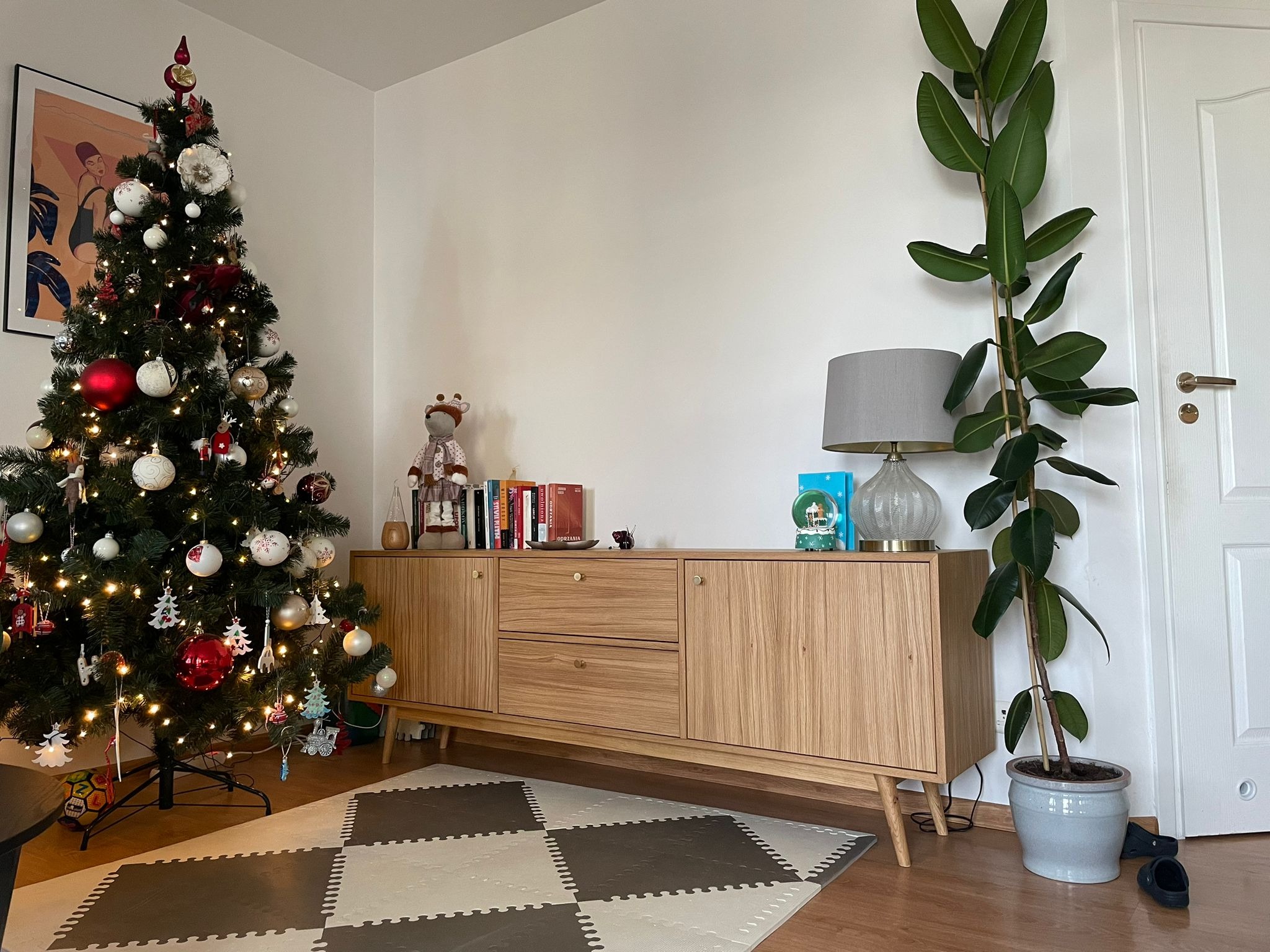 Drewniana komoda w salonie z choinką i rośliną doniczkową. Mebel z jasnego drewna z szufladami i szafkami, na nóżkach. Udekorowane wnętrze w neutralnych barwach.