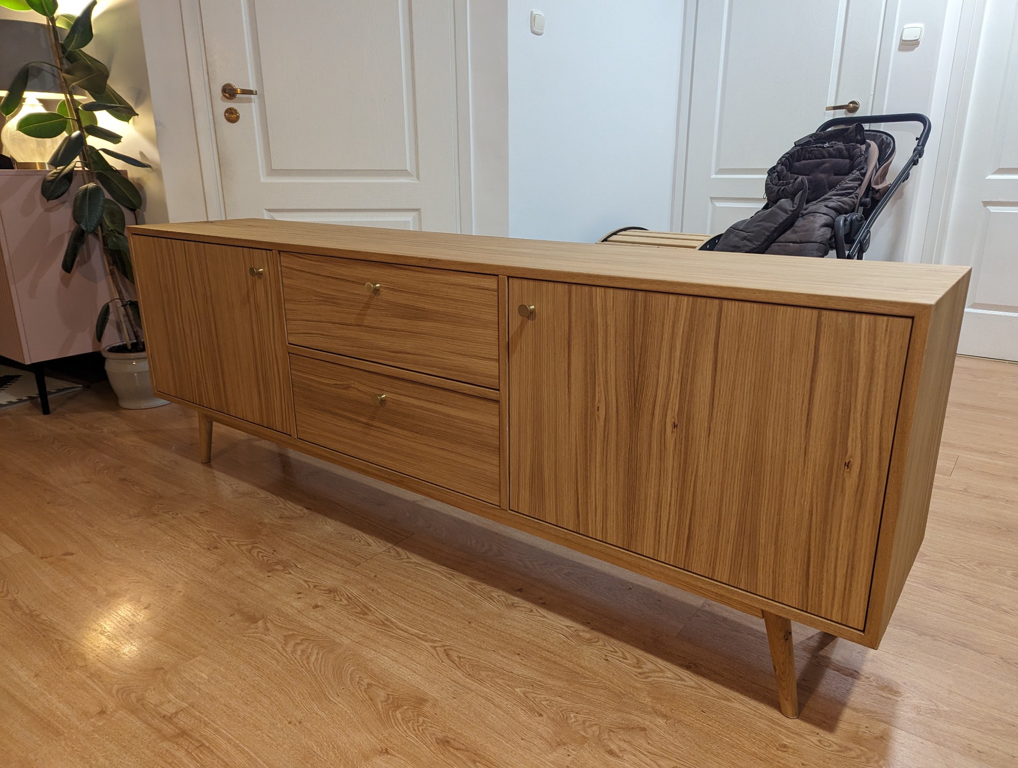 Drewniana komoda z dwiema szufladami i szafkami, widoczna na tle jasnych drzwi i podłogi. Minimalistyczny design, naturalne wykończenie drewna, złote gałki.