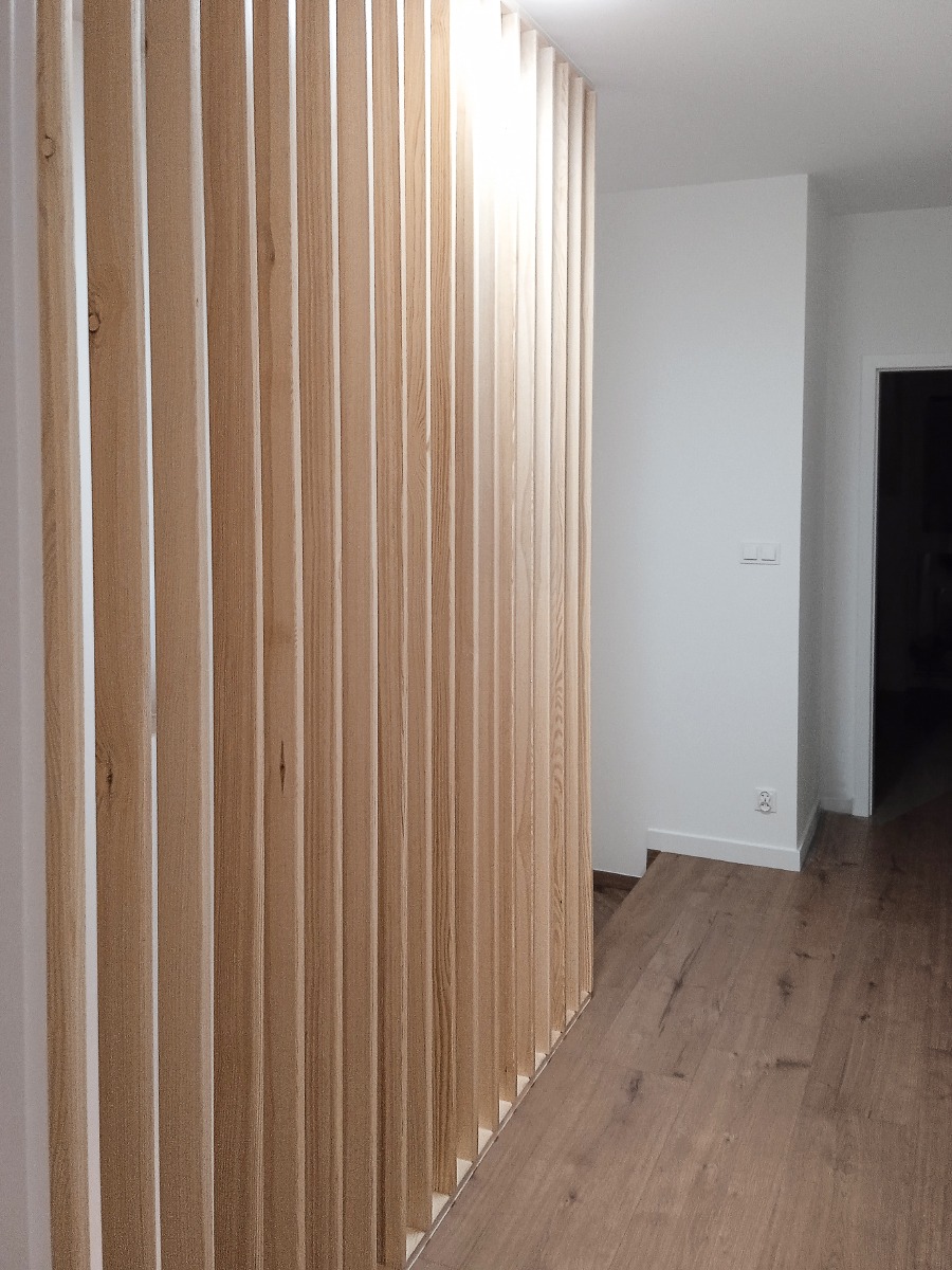 Drewniana ścianka działowa z pionowych listew w jasnym odcieniu drewna, oddzielająca przestrzeń w nowoczesnym wnętrzu z parkietem i białymi ścianami.