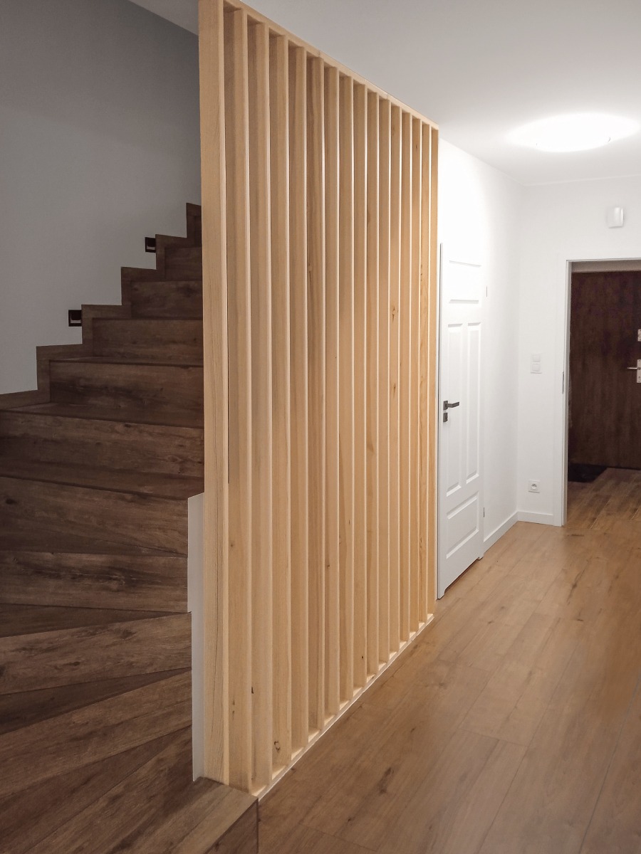 Drewniana ścianka działowa z pionowych listew, oddzielająca schody od korytarza. Naturalne drewno, minimalistyczny design, ciepłe oświetlenie.