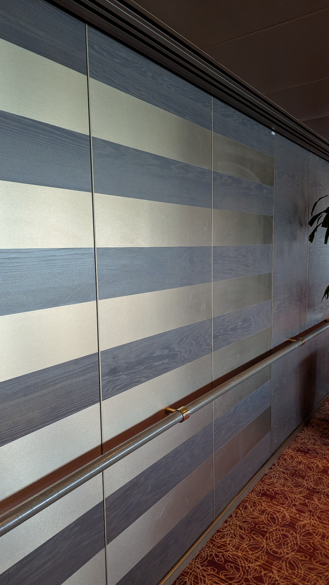 Panel ścienny z poziomymi pasami w odcieniach szarości i srebra, z metalową poręczą. Widok z boku, ukazujący strukturę drewna i połysk metalu.