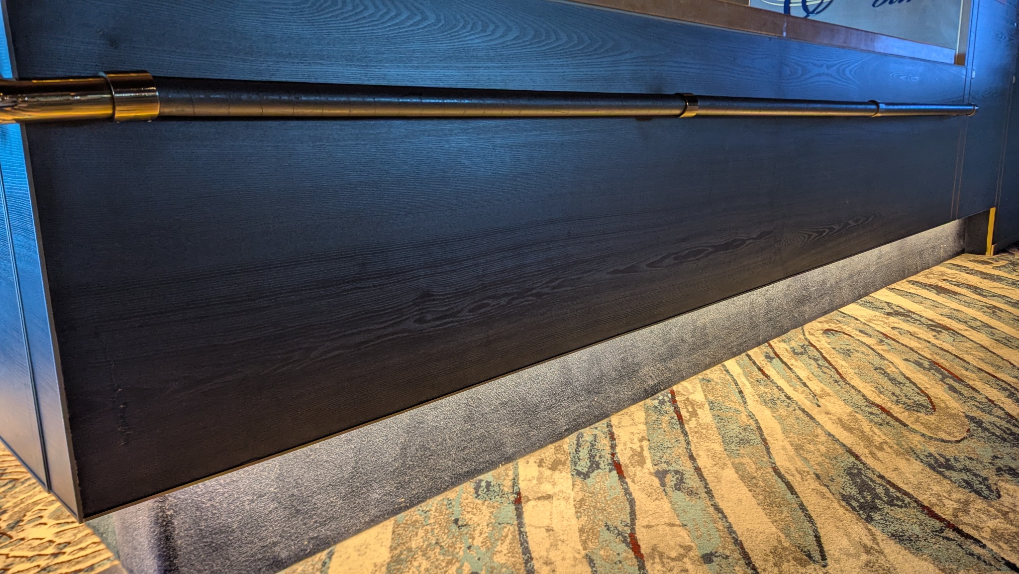 Ciemna, drewniana zabudowa z poziomym, metalowym relingiem i subtelnym oświetleniem przypodłogowym, na tle wzorzystej wykładziny. Detal wykończenia wnętrza.