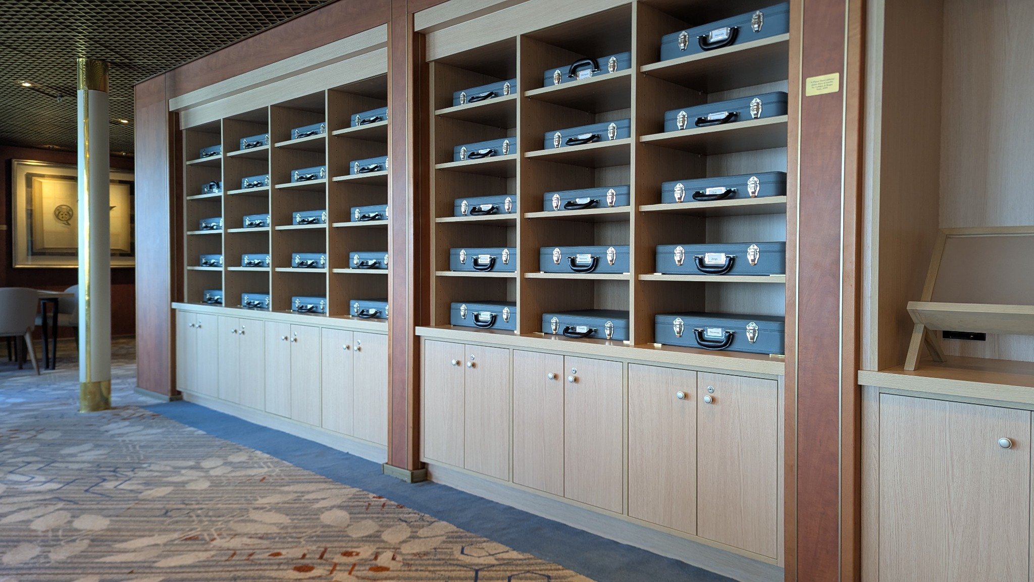 Regał z jasnego drewna z szufladami i półkami, na których ułożone są szare walizki. Widoczny fragment wnętrza z dywanem w geometryczne wzory.