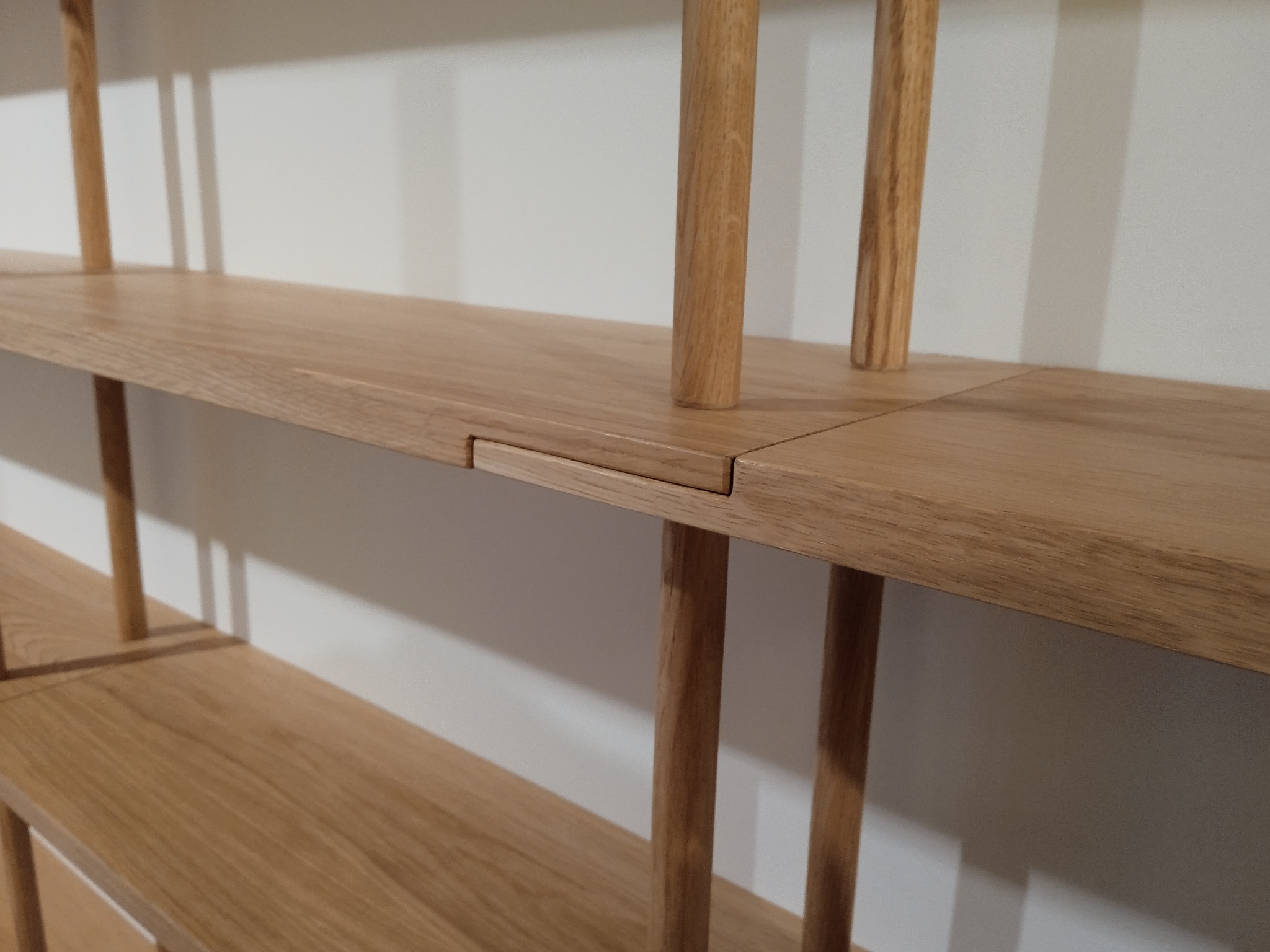 Drewniany regał z jasnego drewna dębowego, minimalistyczny design, widoczne detale połączeń elementów, gładka powierzchnia półek, neutralne tło ściany.