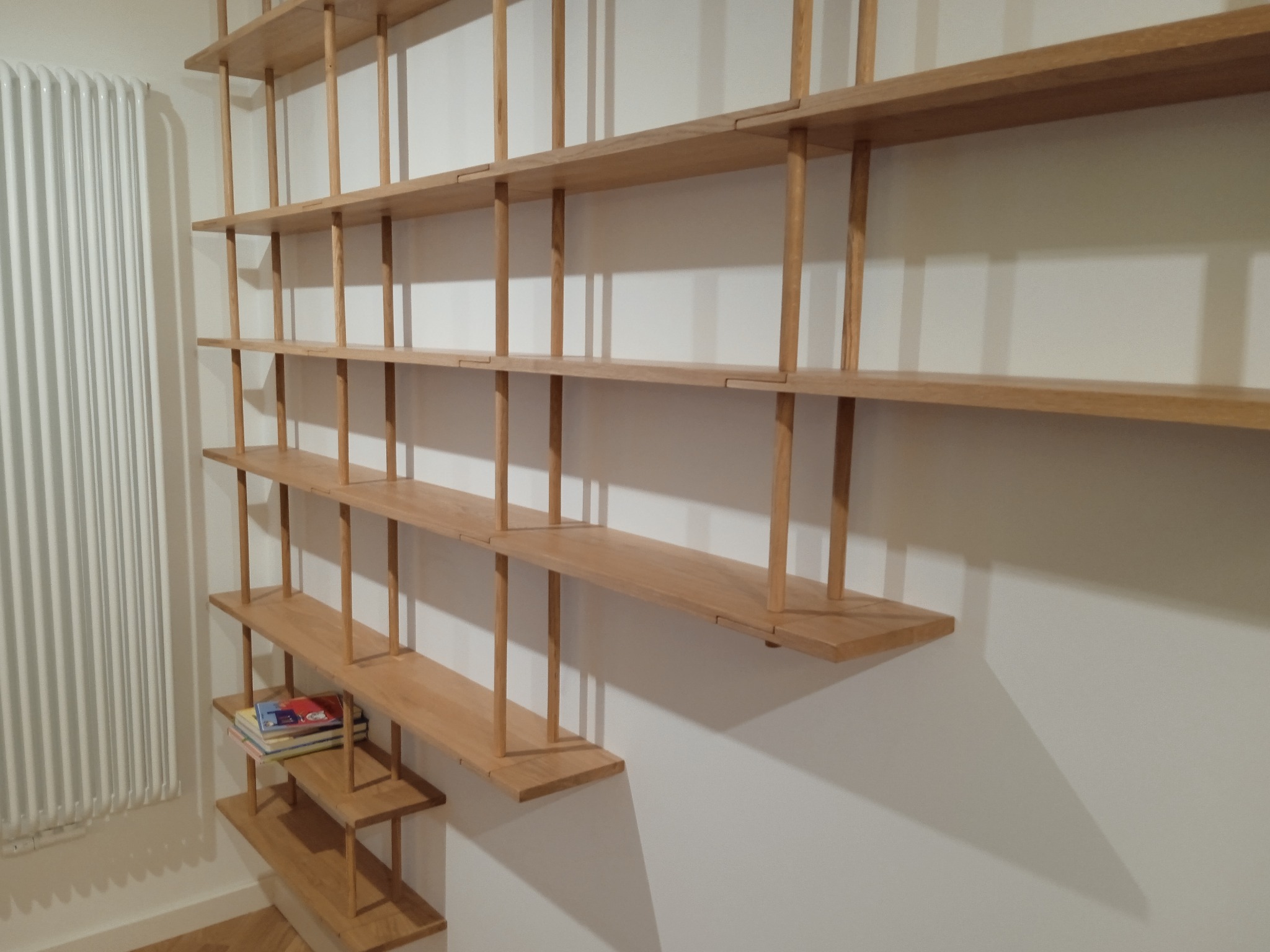 Drewniany regał na książki o minimalistycznym designie, z jasnego drewna, zamontowany na białej ścianie. Na półkach leżą książki. Widoczny grzejnik.
