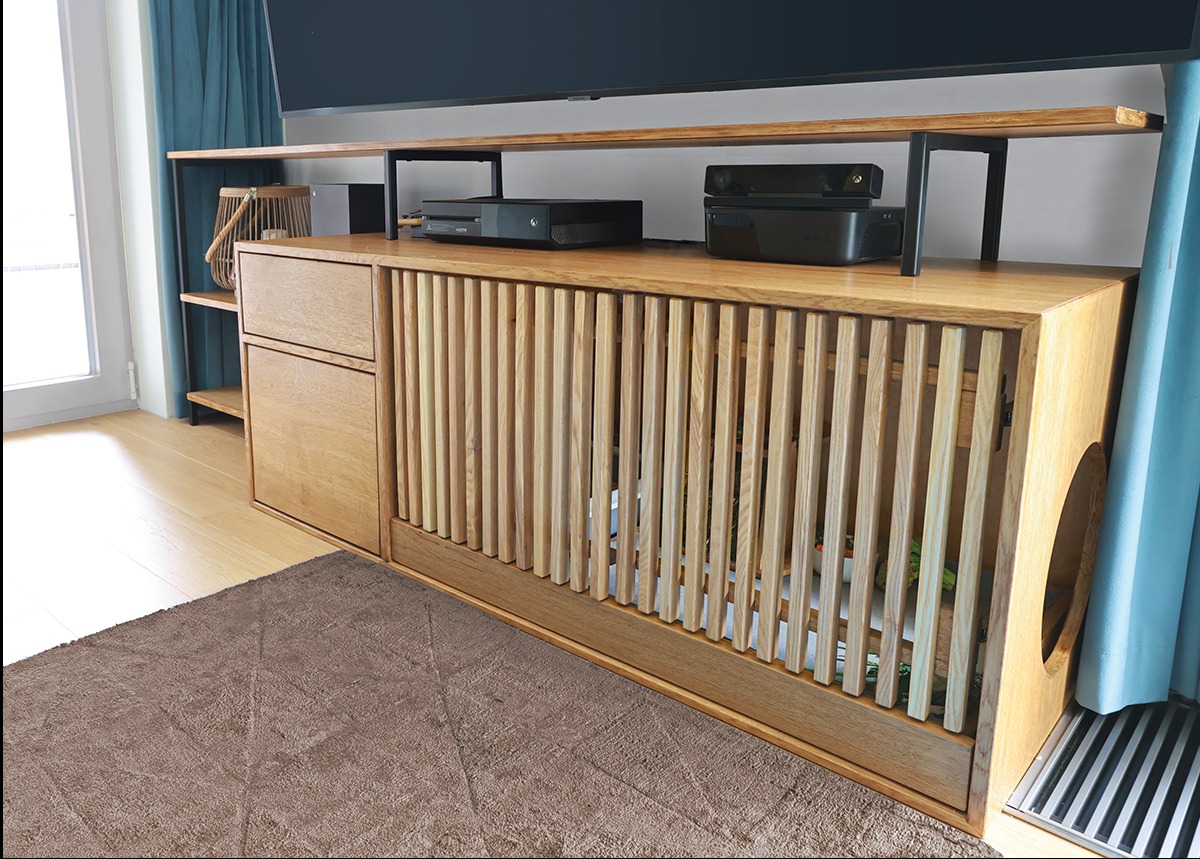 Drewniana szafka RTV z lamelami i półkami, na której stoi sprzęt elektroniczny. Widoczny fragment okna z zasłoną i dywan. Unikalny design z otworem na boku.