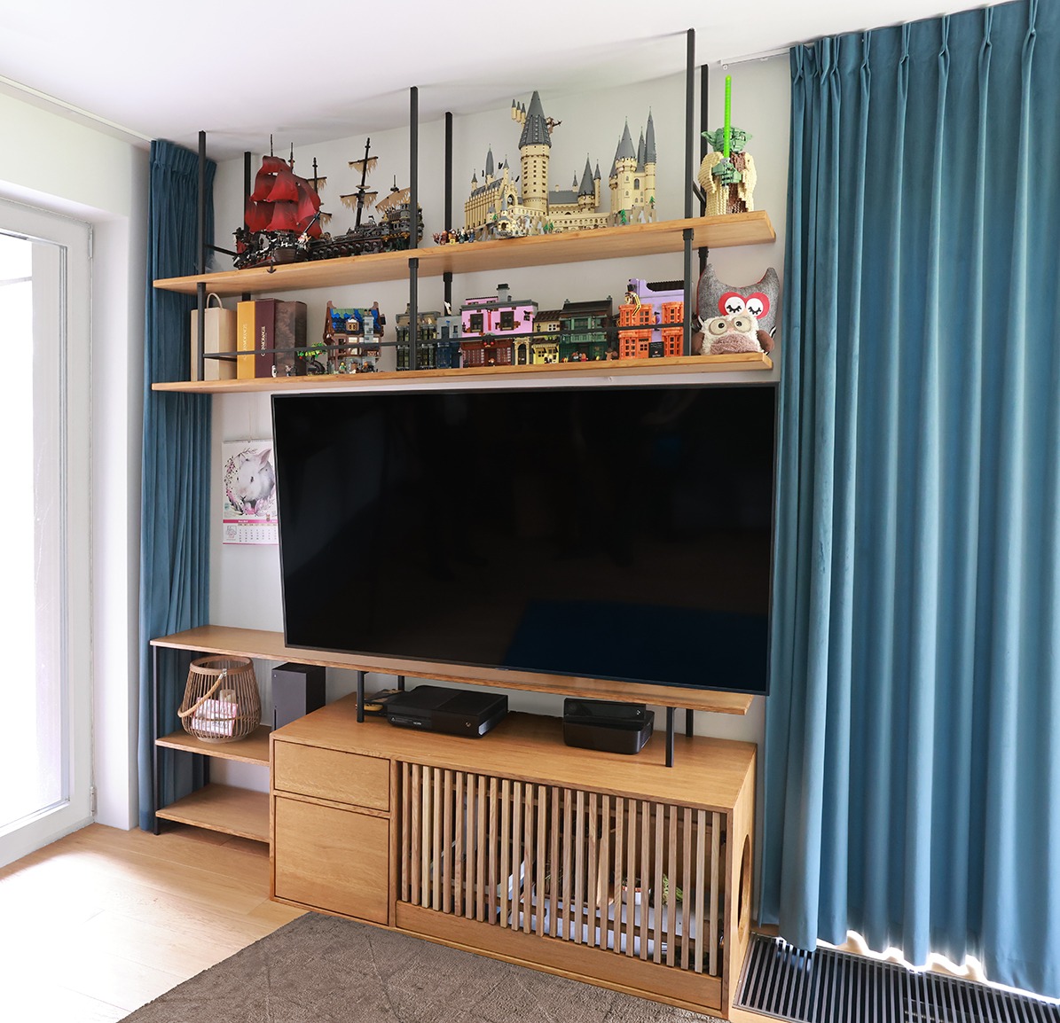 Drewniana szafka RTV z lamelami i półkami pod sufitem, na których stoją modele budowli z klocków. Całość w jasnym, nowoczesnym wnętrzu z błękitnymi zasłonami.