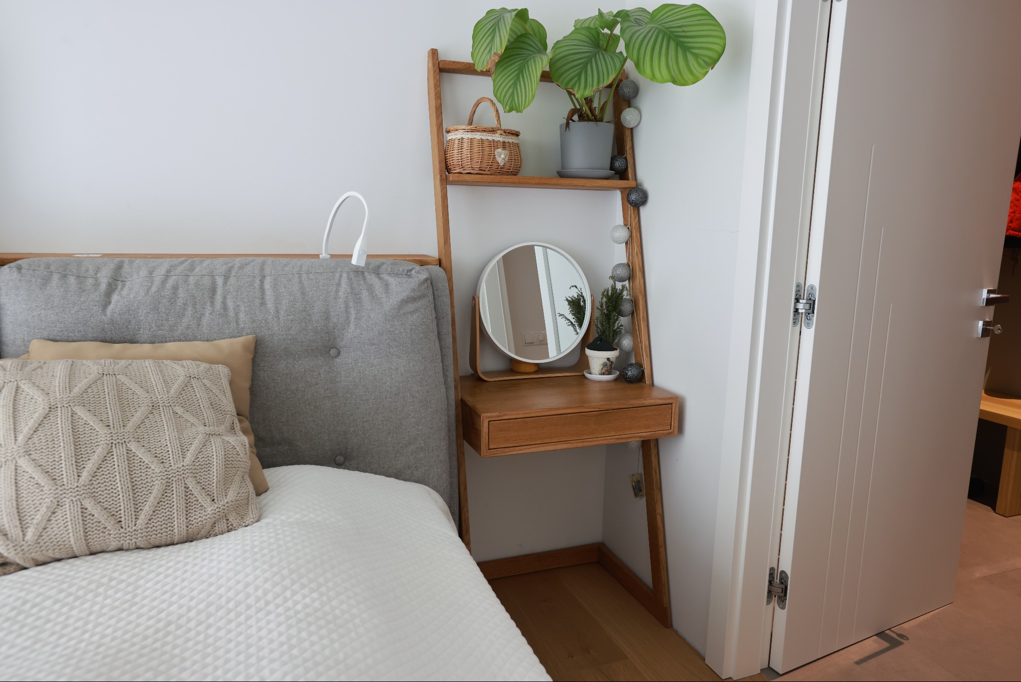 Drewniana drabina-regał z lustrem i szufladą, ustawiona przy łóżku z szarym zagłówkiem. Na półkach koszyk, roślina w doniczce i dekoracje. Białe ściany i drzwi.