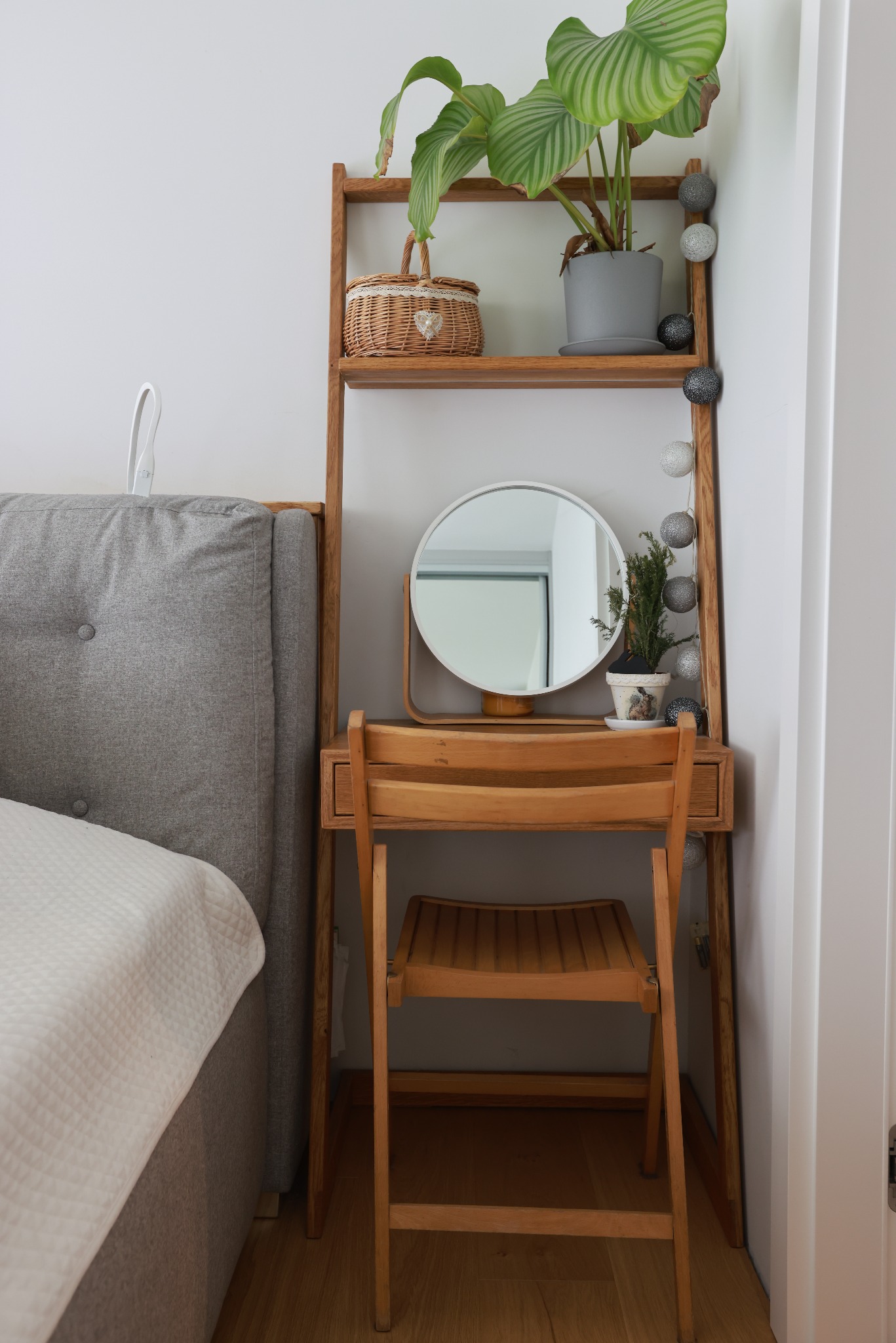Drewniana toaletka z okrągłym lustrem i krzesłem w jasnym wnętrzu. Na półkach dekoracje: roślina w doniczce, koszyk i girlanda świetlna. Obok wezgłowie łóżka.