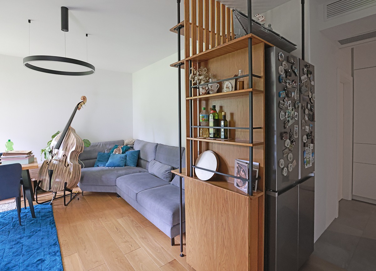 Nowoczesny salon z drewnianą zabudową regałową, dzielącą przestrzeń. Widoczny szary narożnik, kontrabas i okrągła lampa. Parkiet z naturalnego drewna.