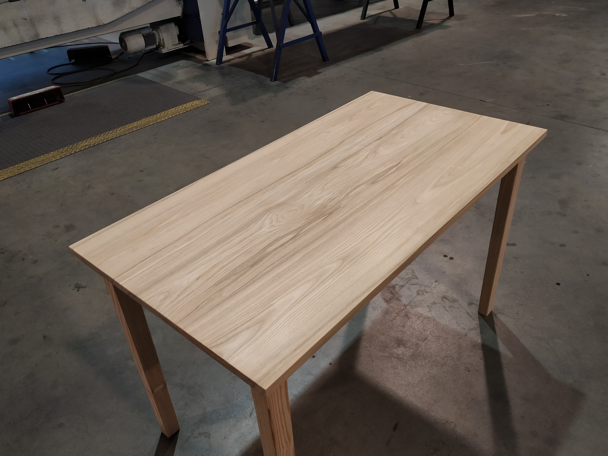 Drewniany stół z jasnego drewna w warsztacie w Gdańsku. Minimalistyczny design, widoczne słoje drewna. Stół na czterech nogach, surowy beton na podłodze.