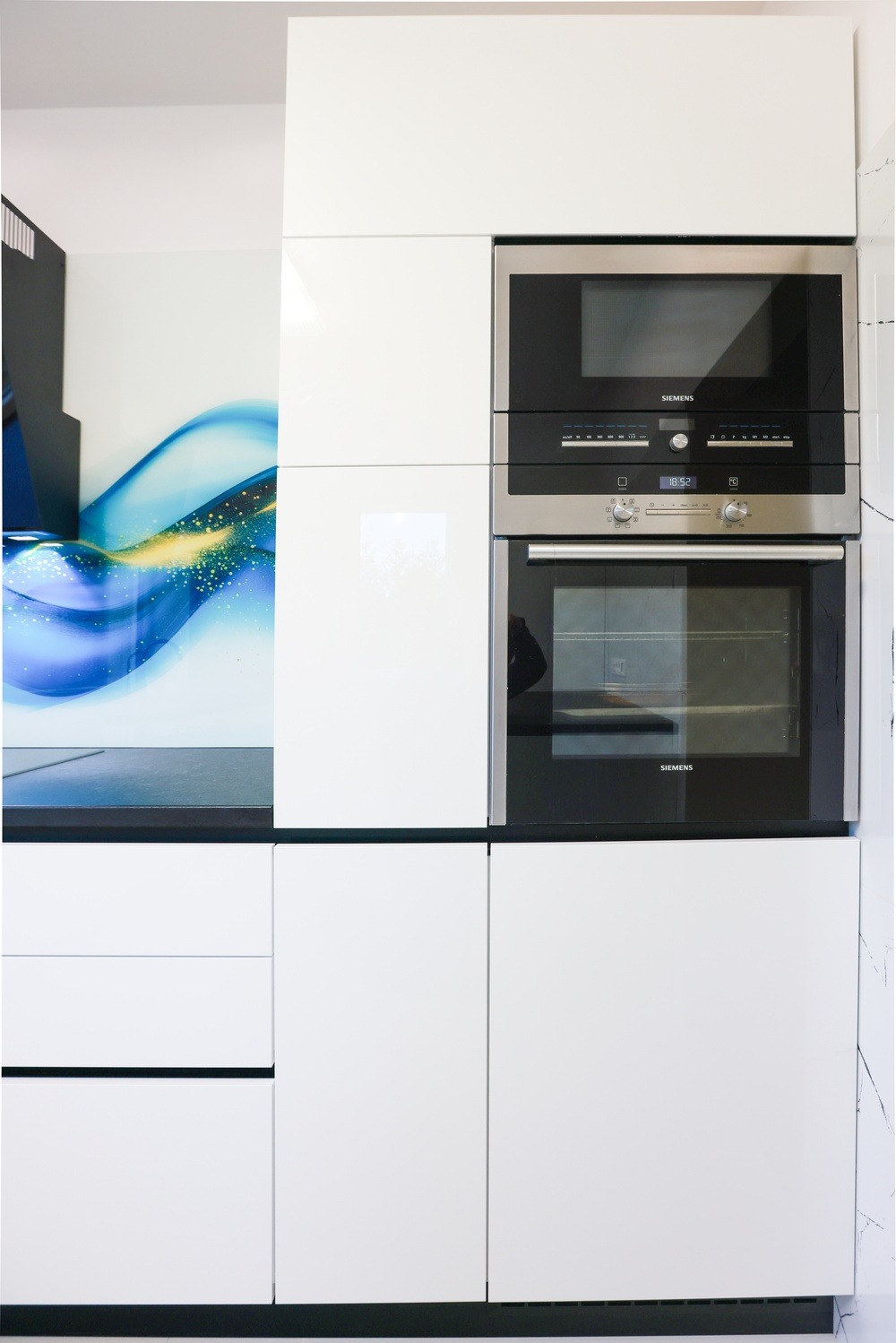 Biała zabudowa kuchenna z piekarnikiem i mikrofalówką Siemens, fronty lakierowane na wysoki połysk, dekoracyjny panel szklany z motywem abstrakcyjnym.