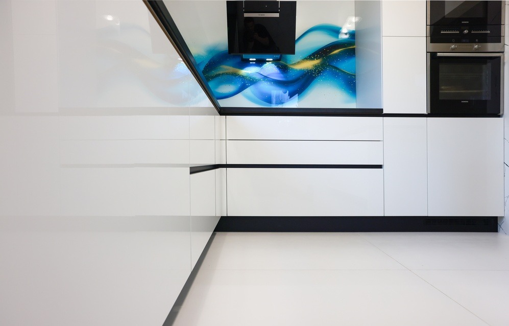 Biała, nowoczesna kuchnia z połyskiem, minimalistyczne fronty szafek, czarne akcenty, abstrakcyjny panel nad blatem, wbudowane sprzęty AGD.