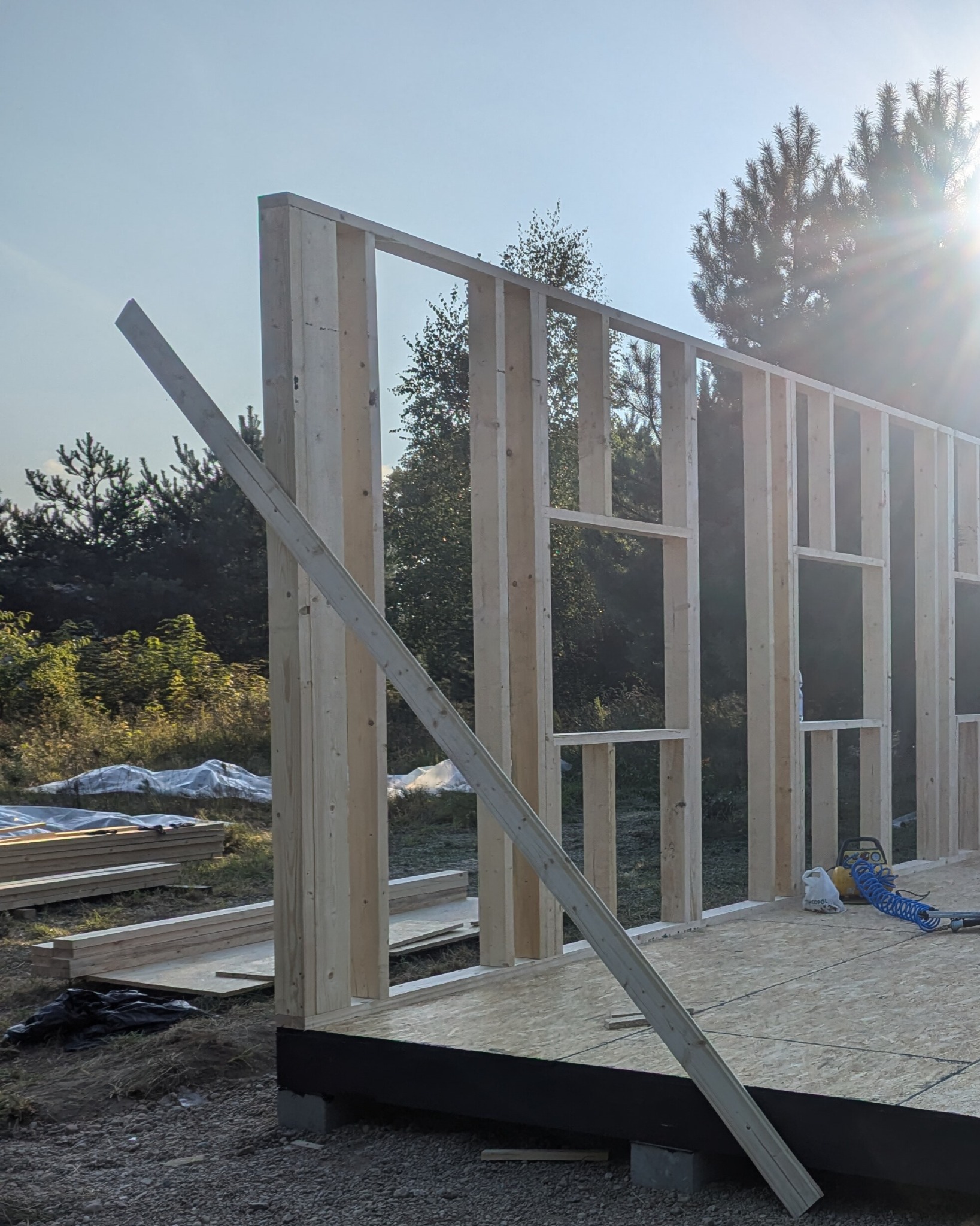 Wznoszenie szkieletu domu: drewniana konstrukcja ściany z otworami okiennymi, podparta deską, narzędzia pneumatyczne obok. Widok na zewnątrz, w tle drzewa.