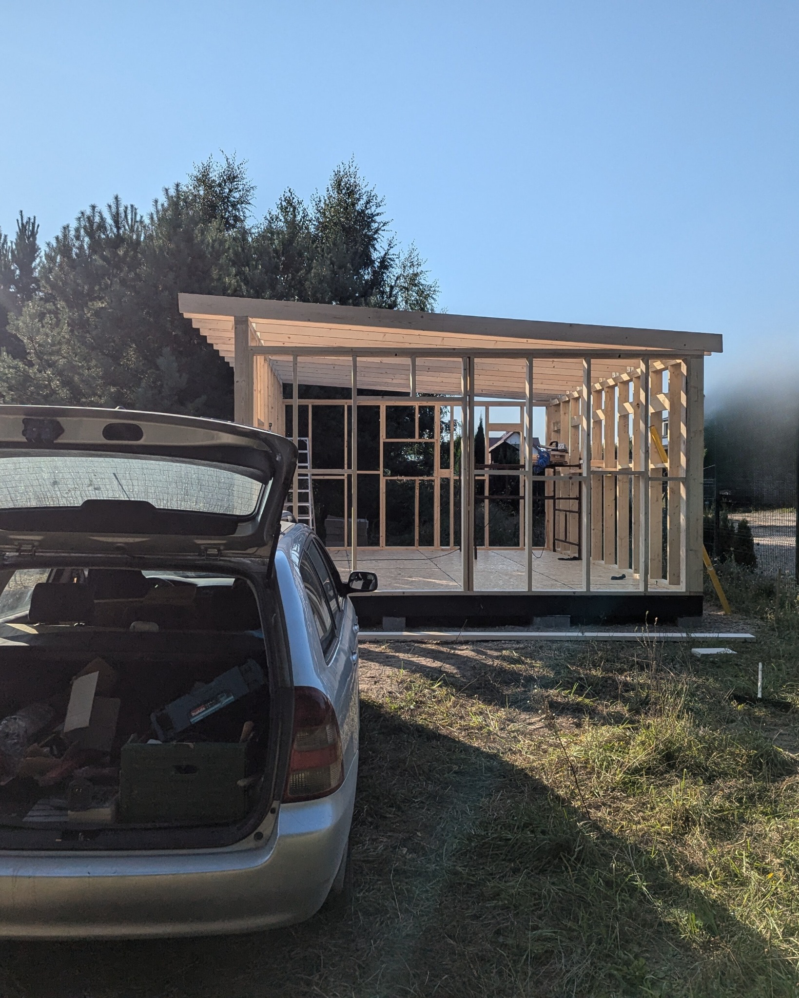 Wznoszenie konstrukcji szkieletowej budynku w ogrodzie, widok zza otwartego bagażnika samochodu z narzędziami. W tle drzewa i błękitne niebo.