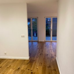 Filip Kierzkowski - Jasne, drewniane panele podłogowe w nowym mieszkaniu, widok z korytarza na balkon przez przeszklone drzwi. Minimalistyczne wnętrze z białymi ścianami i punktowym oświetleniem.
