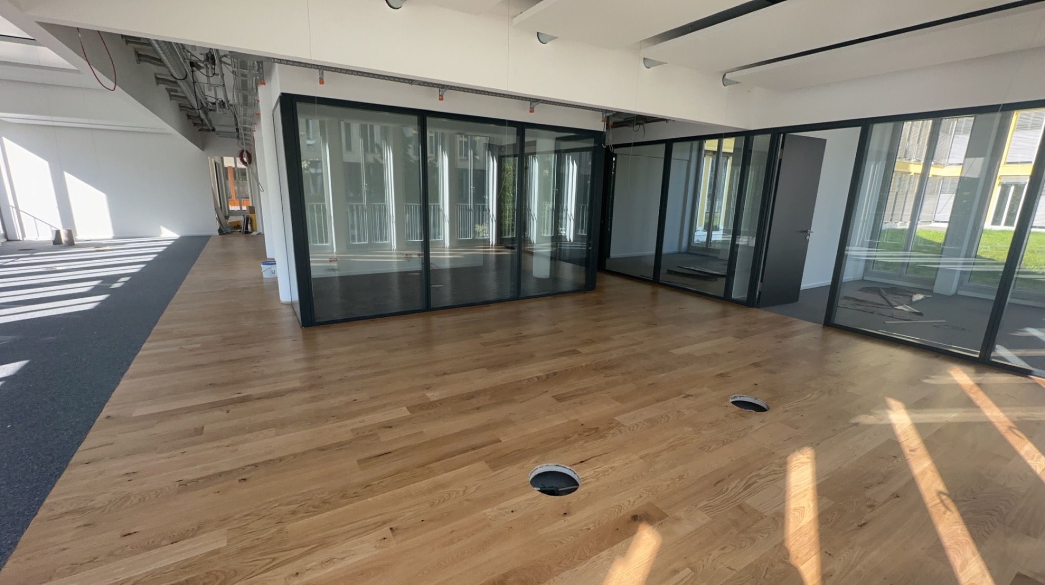 Jasna, drewniana podłoga w biurze z przeszklonymi ścianami działowymi. Widoczne otwory w podłodze. Część podłogi pokryta szarą wykładziną. Oświetlenie sufitowe.