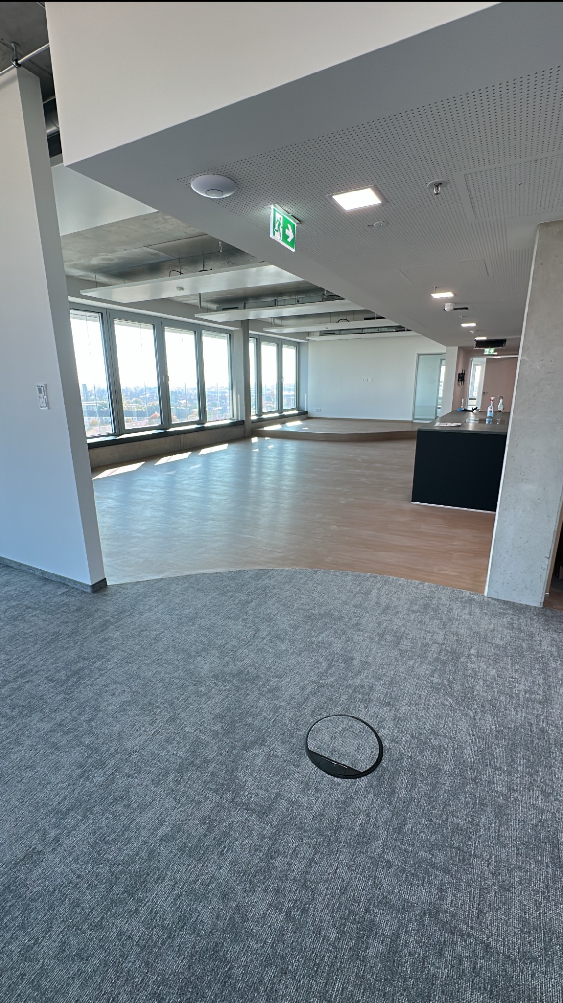 Jasne biuro po remoncie: parkiet i wykładzina dywanowa. Widok na miasto z wysokich okien. Otwór w podłodze na pierwszym planie. Nowoczesne oświetlenie sufitowe.