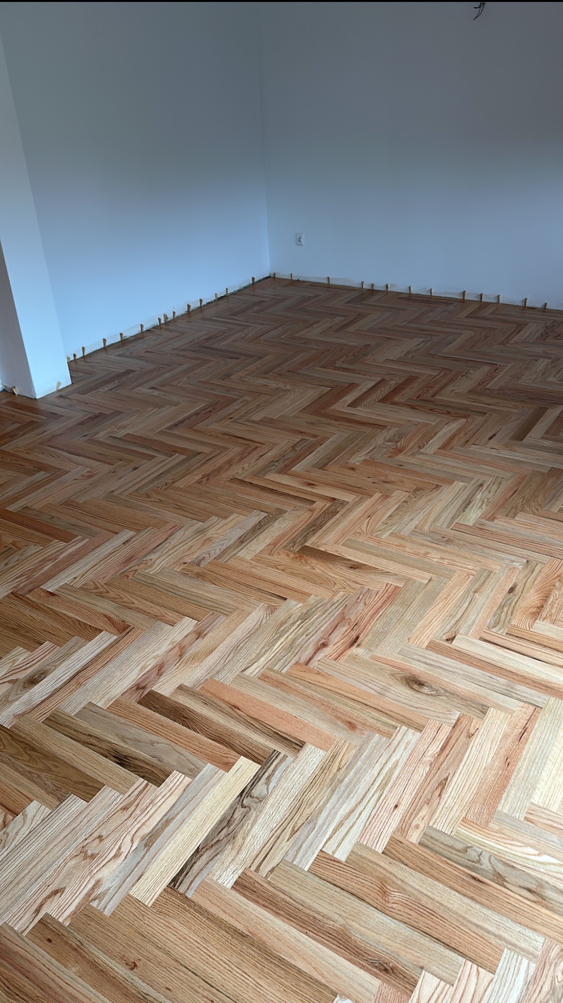 Ułożony parkiet w jodełkę w jasnym pomieszczeniu z białymi ścianami. Widoczne drewniane kliny dystansowe przy ścianach. Naturalne oświetlenie podkreśla wzór drewna.