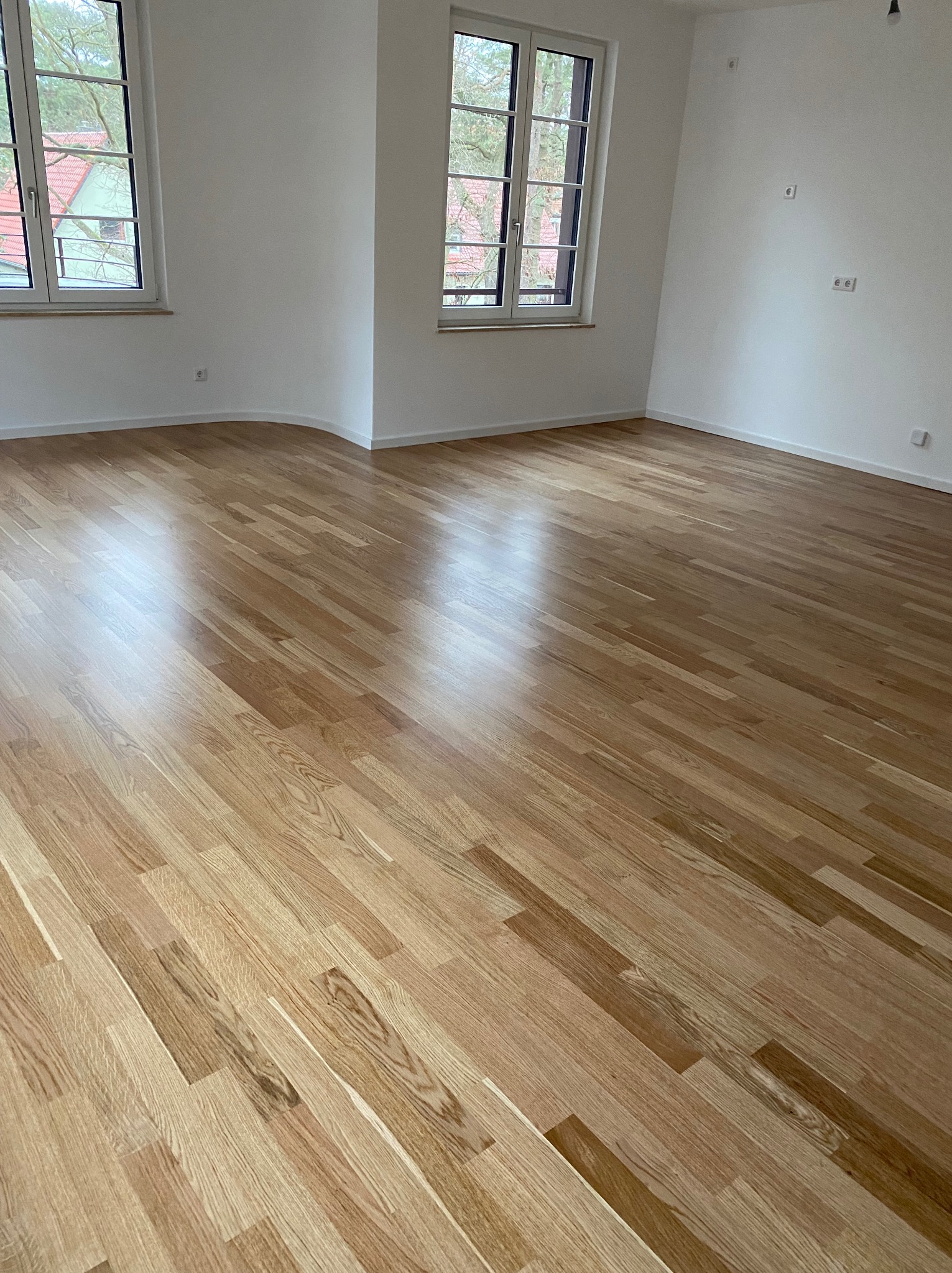 Jasny, drewniany parkiet ułożony w pustym pokoju z dwoma oknami. Widoczne gniazdka elektryczne na białych ścianach. Naturalne światło podkreśla wzór drewna.