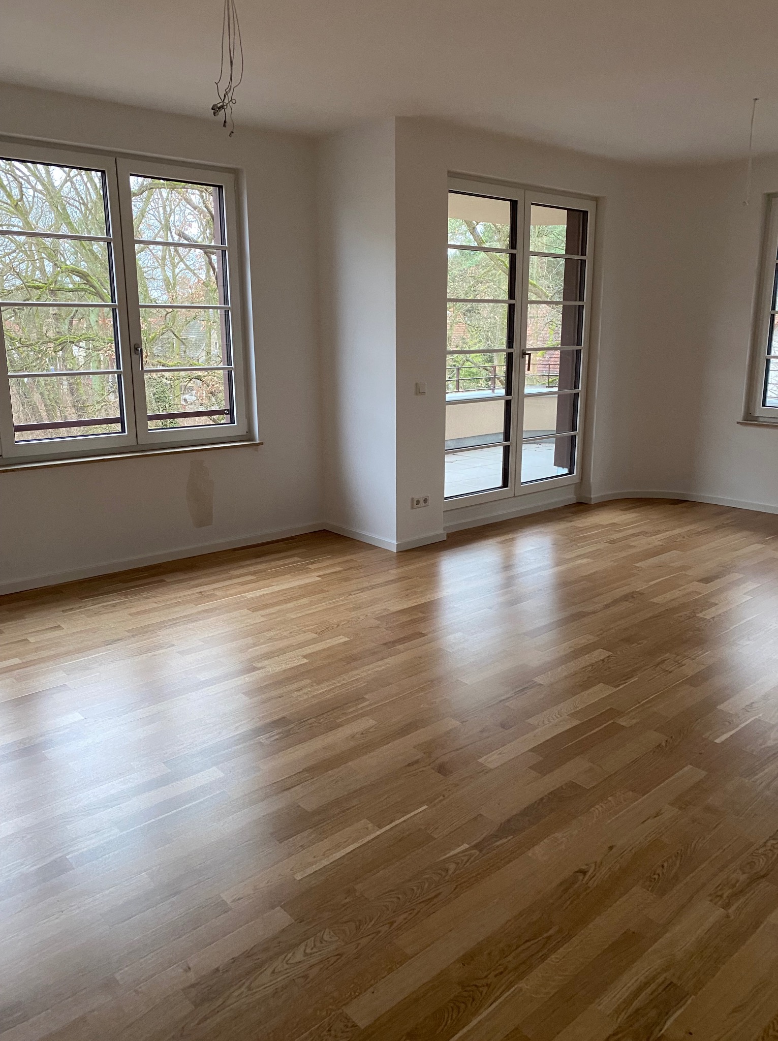 Jasny parkiet w pustym pokoju z oknami i drzwiami balkonowymi. Widoczne ślady po remoncie na ścianie. Naturalne światło podkreśla strukturę drewna.