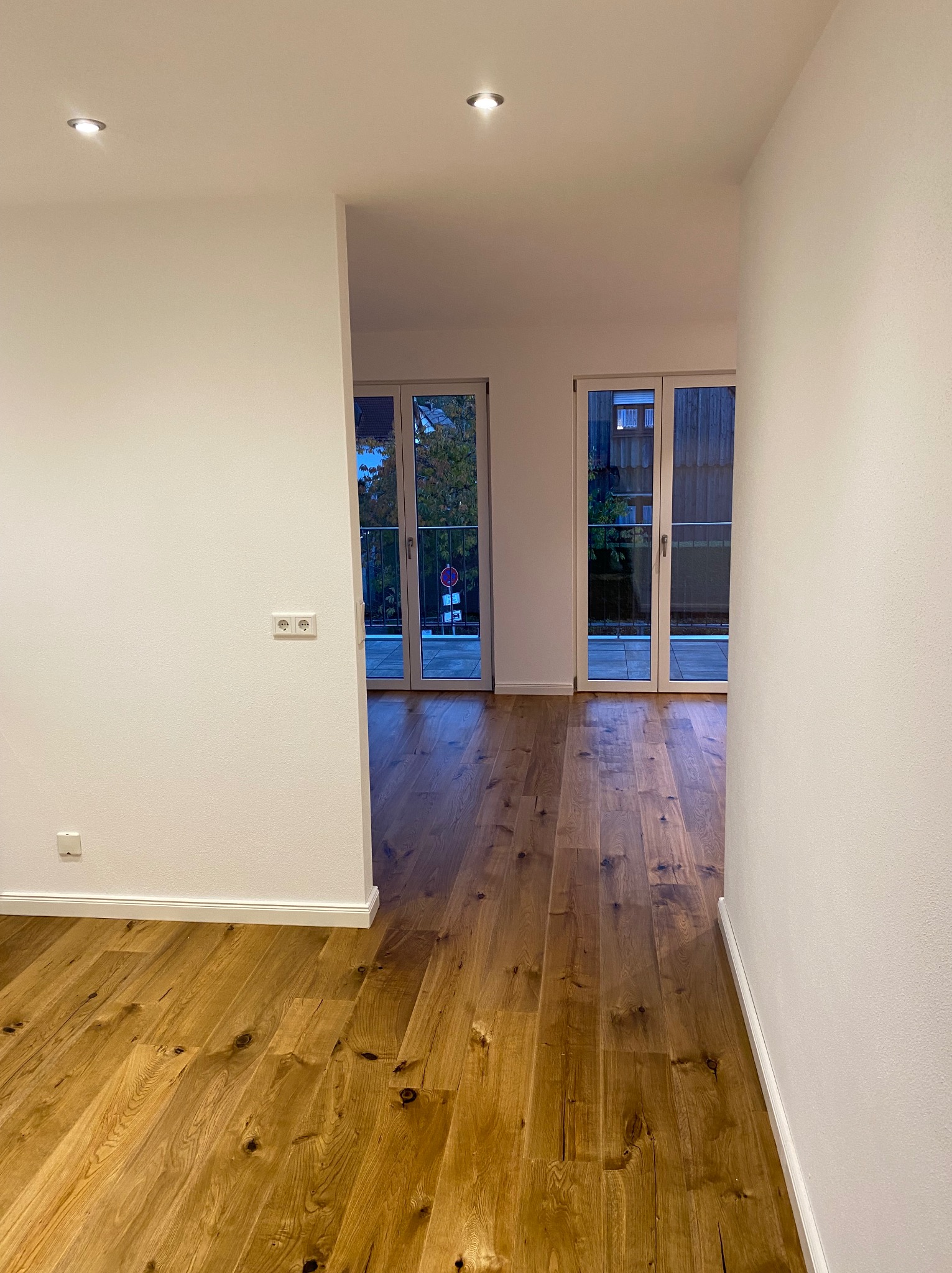 Jasne, drewniane panele podłogowe w nowym mieszkaniu, widok z korytarza na balkon przez przeszklone drzwi. Minimalistyczne wnętrze z białymi ścianami i punktowym oświetleniem.