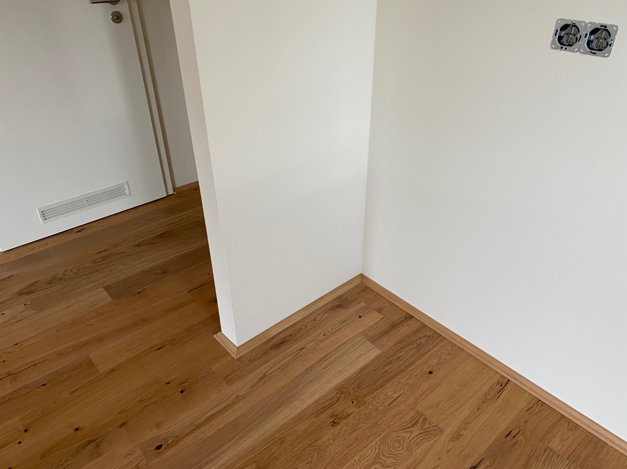 Jasny parkiet z desek w nowym wnętrzu, białe ściany z gniazdkami elektrycznymi, listwy przypodłogowe. Widok na drzwi.