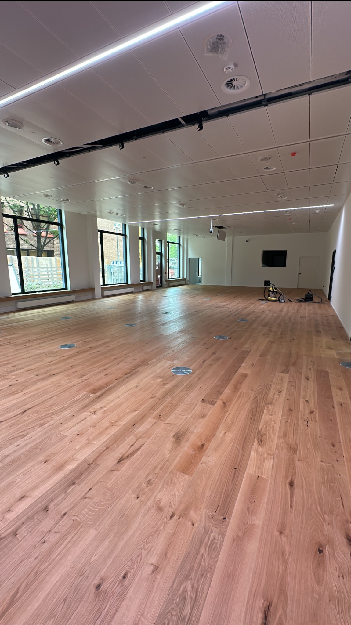 Jasna, przestronna sala z nową, drewnianą podłogą. Widoczne okna z widokiem na zieleń, białe ściany i sufit z oświetleniem. Na końcu sali odkurzacz przemysłowy.