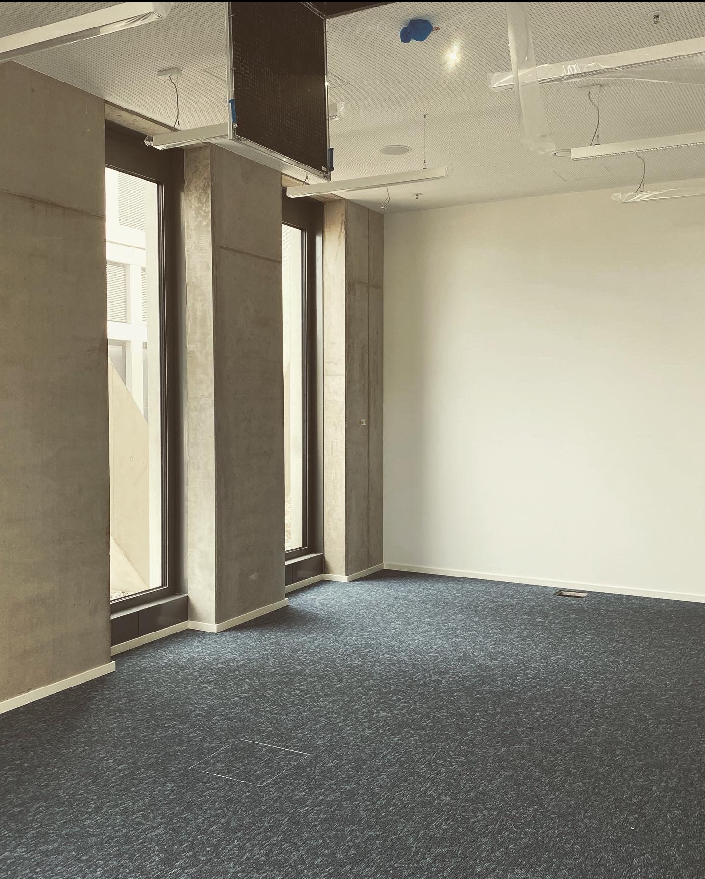 Pusta przestrzeń biurowa z szarą wykładziną, betonowymi filarami okiennymi i białymi ścianami. Widoczne elementy instalacji pod sufitem. Surowe wykończenie, minimalistyczny styl.