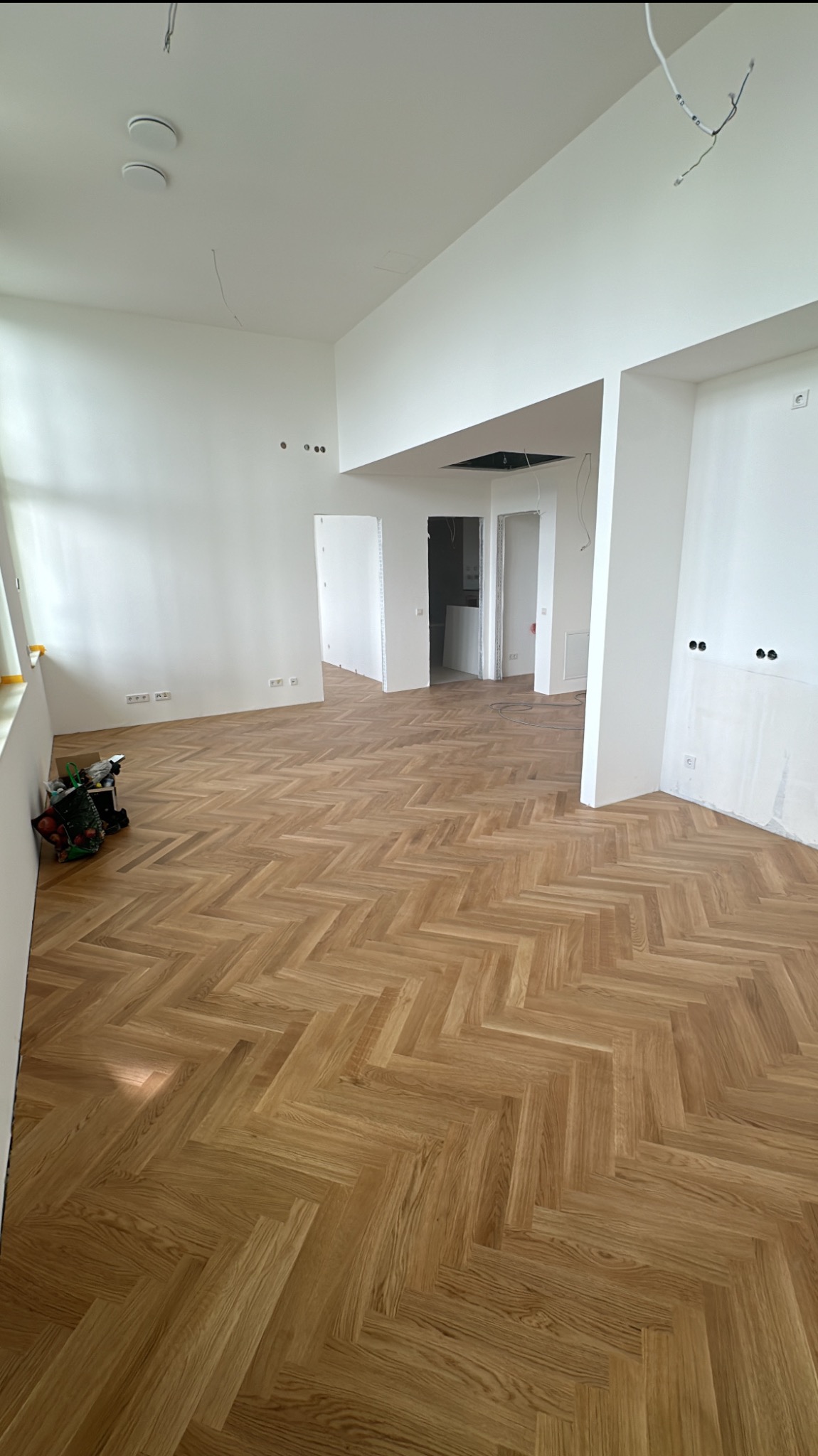 Jasny parkiet ułożony we wzór jodełki francuskiej w przestronnym, odświeżonym wnętrzu. Widoczne narzędzia w rogu, surowe ściany i otwarte przejścia do innych pomieszczeń.