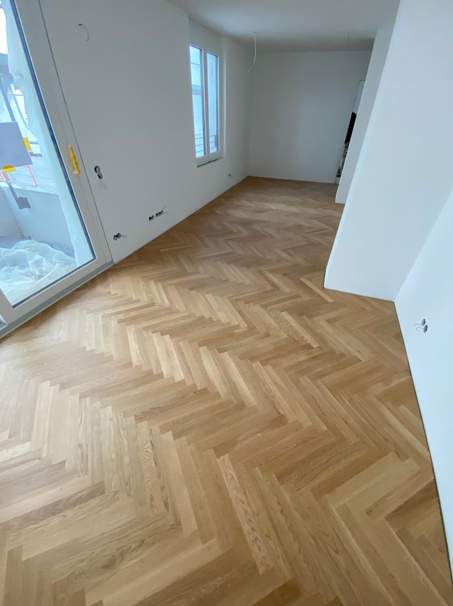 Jasny parkiet ułożony w jodełkę w nowym, pustym pokoju z białymi ścianami i oknem. Widoczne gniazdka elektryczne i fragment okna balkonowego.