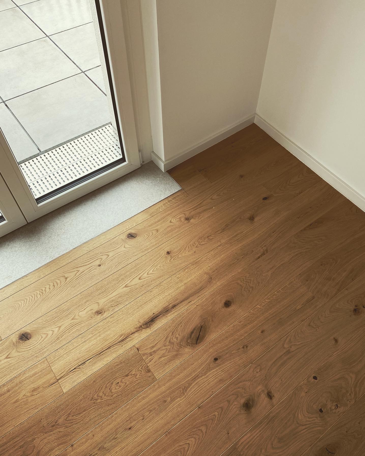 Jasna, drewniana podłoga z wyraźnym usłojeniem, ułożona wzdłuż ściany z białą listwą przypodłogową. Widoczny fragment balkonu z szarą terakotą i kratką odpływową.