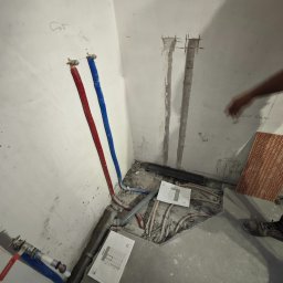 MJM SPÓŁKA Z OGRANICZONĄ ODPOWIEDZIALNOŚCIĄ - Wnętrze łazienki w trakcie prac instalacyjnych: widoczne rury, bruzdy w ścianie, płytki i plany. Etap przygotowawczy przed wykończeniem.