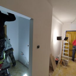 MJM SPÓŁKA Z OGRANICZONĄ ODPOWIEDZIALNOŚCIĄ - Wnętrze w trakcie prac wykończeniowych: widoczne ściany po szpachlowaniu, instalacje elektryczne i hydrauliczne, drabina i lampa budowlana. Dwóch pracowników w trakcie pracy.