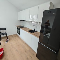 MJM SPÓŁKA Z OGRANICZONĄ ODPOWIEDZIALNOŚCIĄ - Nowoczesna, jasna kuchnia z białymi szafkami i drewnianym blatem. Widoczny czarny zlew, płyta grzewcza i lodówka. Na podłodze mop i wiadro, co sugeruje sprzątanie po remoncie.