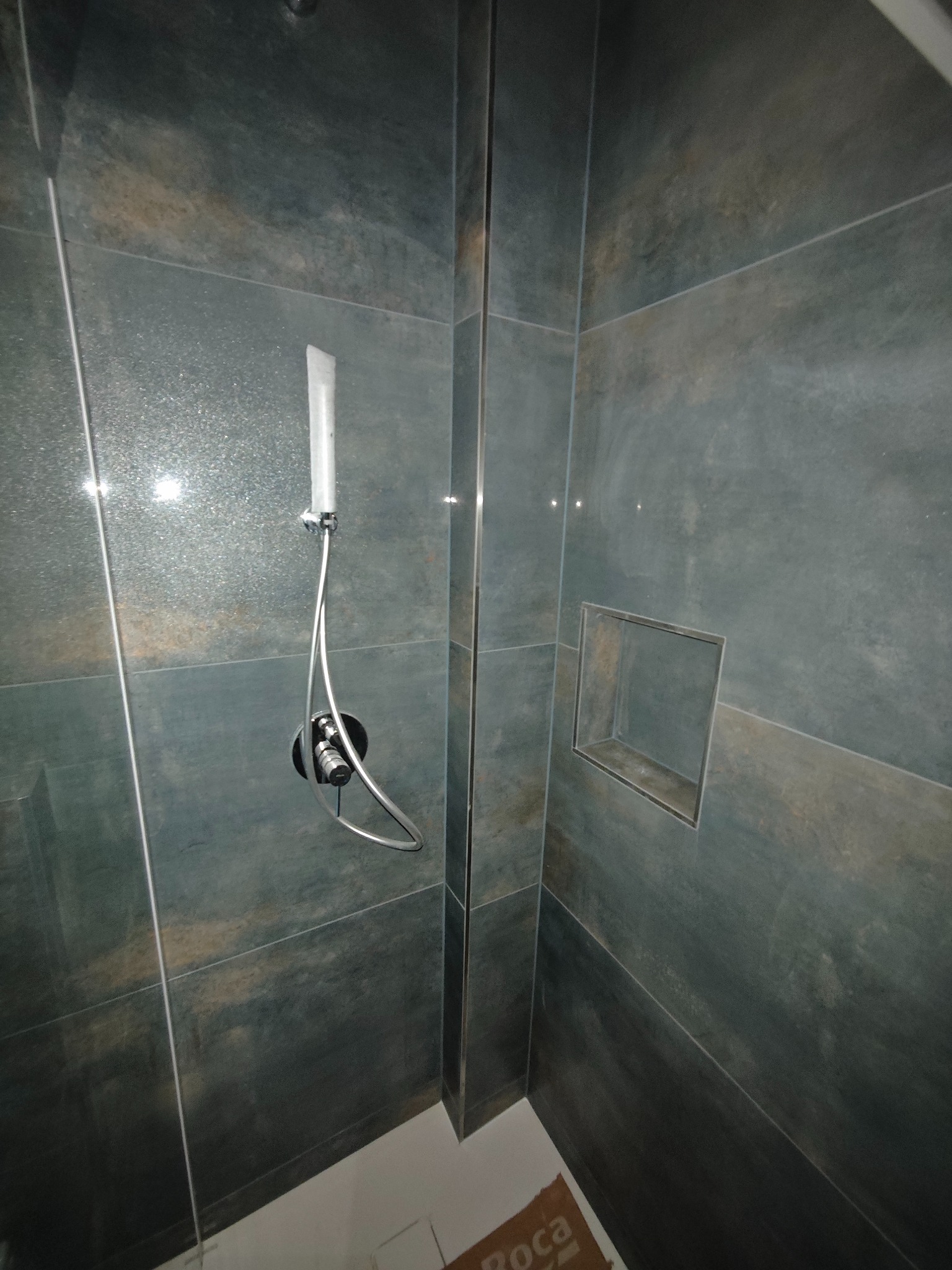 Kabina prysznicowa z szarymi płytkami imitującymi kamień, wnęka w ścianie, deszczownica z czarną baterią. Widoczny fragment podłogi i logo Roca.