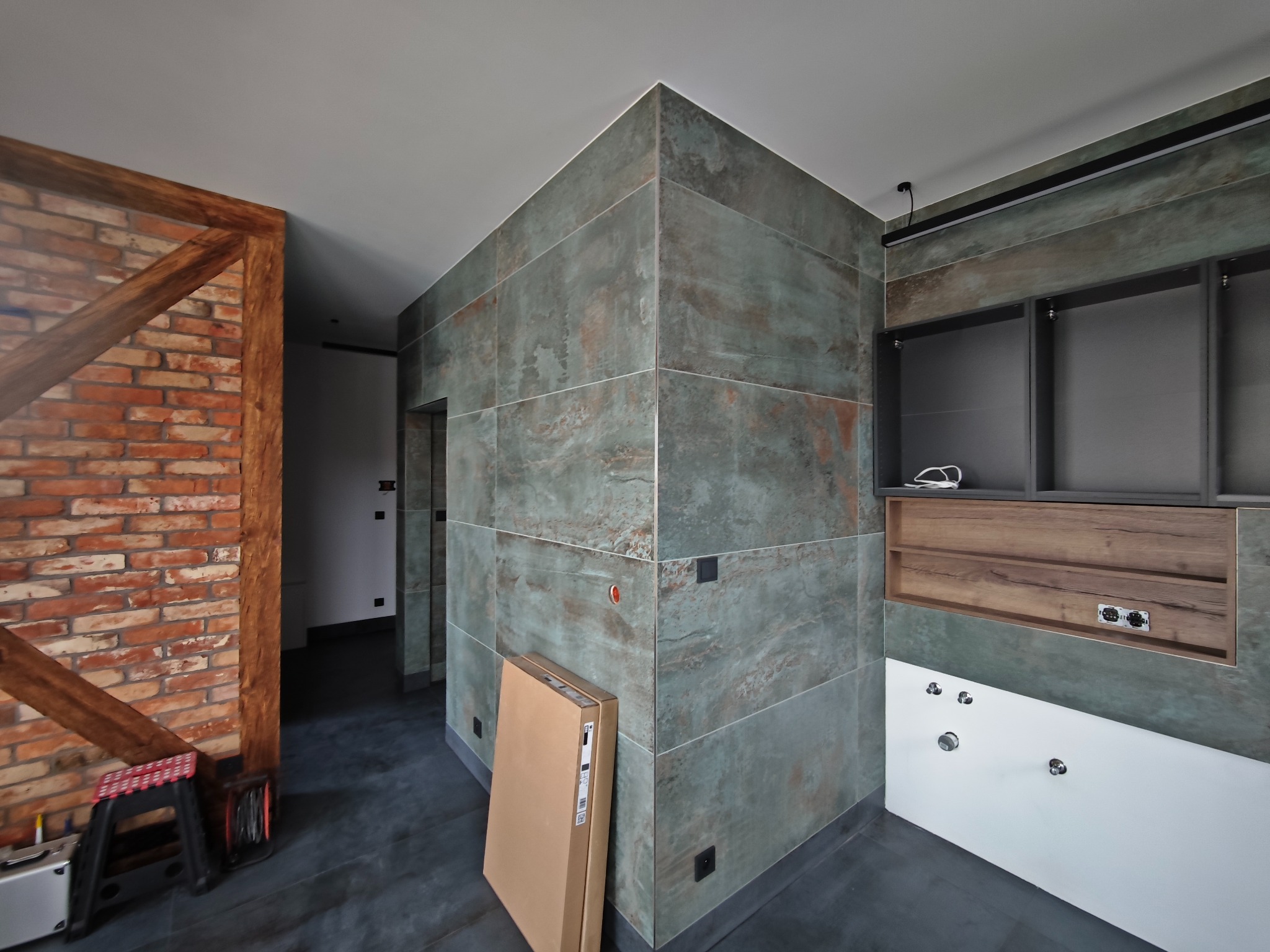 Nowoczesna łazienka w trakcie wykańczania: zielono-szare płytki, drewniane akcenty, widoczne instalacje i karton, podkreślające etap prac wykończeniowych.