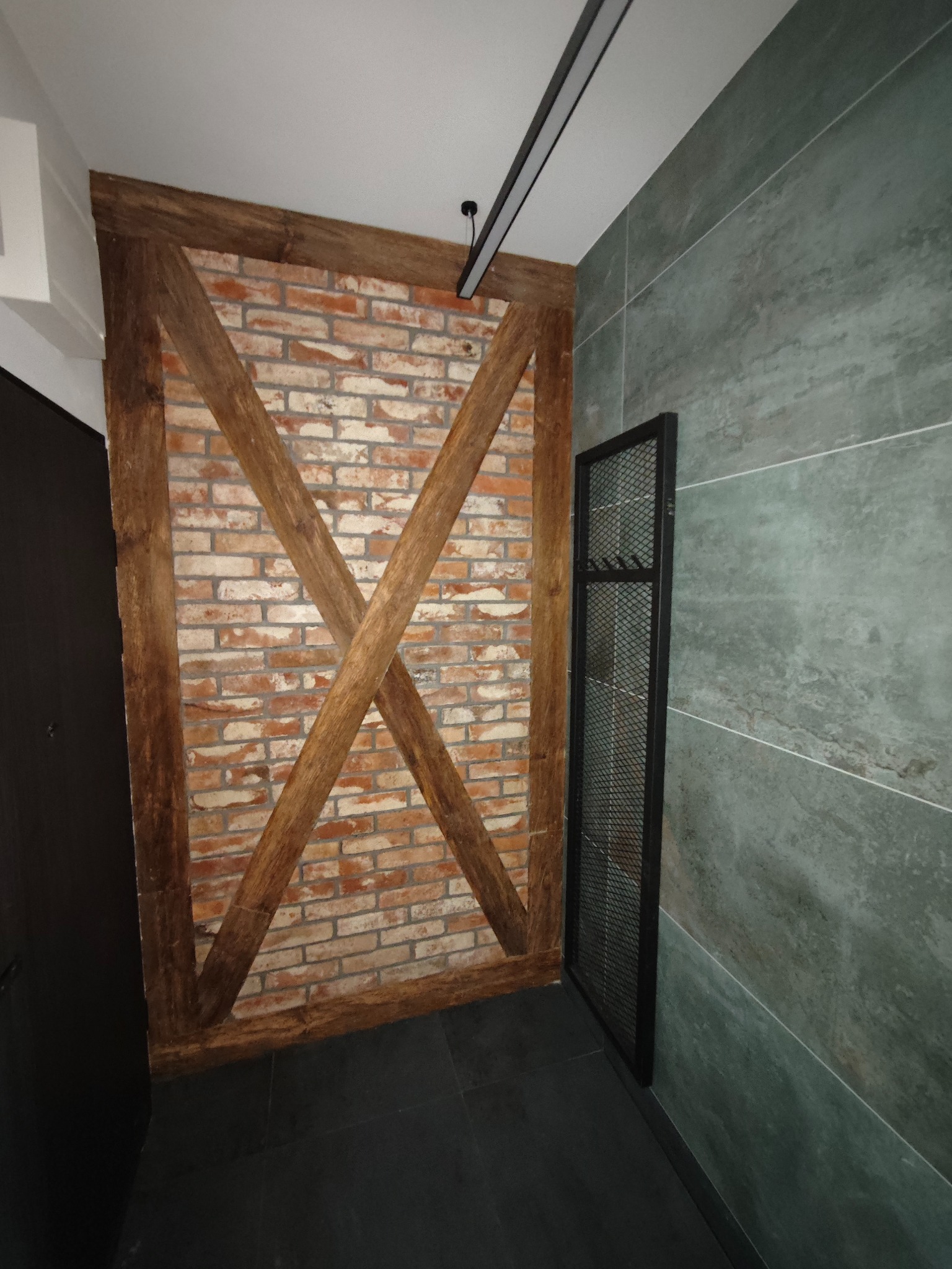 Ceglana ściana z drewnianą ramą w kształcie litery X, szara ściana z płytek i czarna podłoga tworzą nowoczesne wnętrze. Minimalistyczny design z industrialnym akcentem.