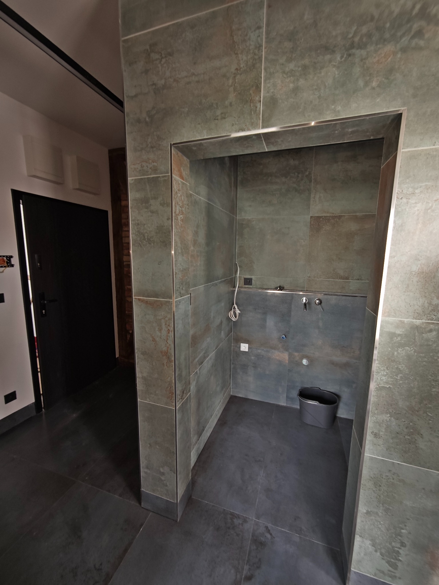 Nowoczesna łazienka z szarymi płytkami imitującymi kamień, widoczna umywalka i wiadro. Minimalistyczny design z akcentami metalu.