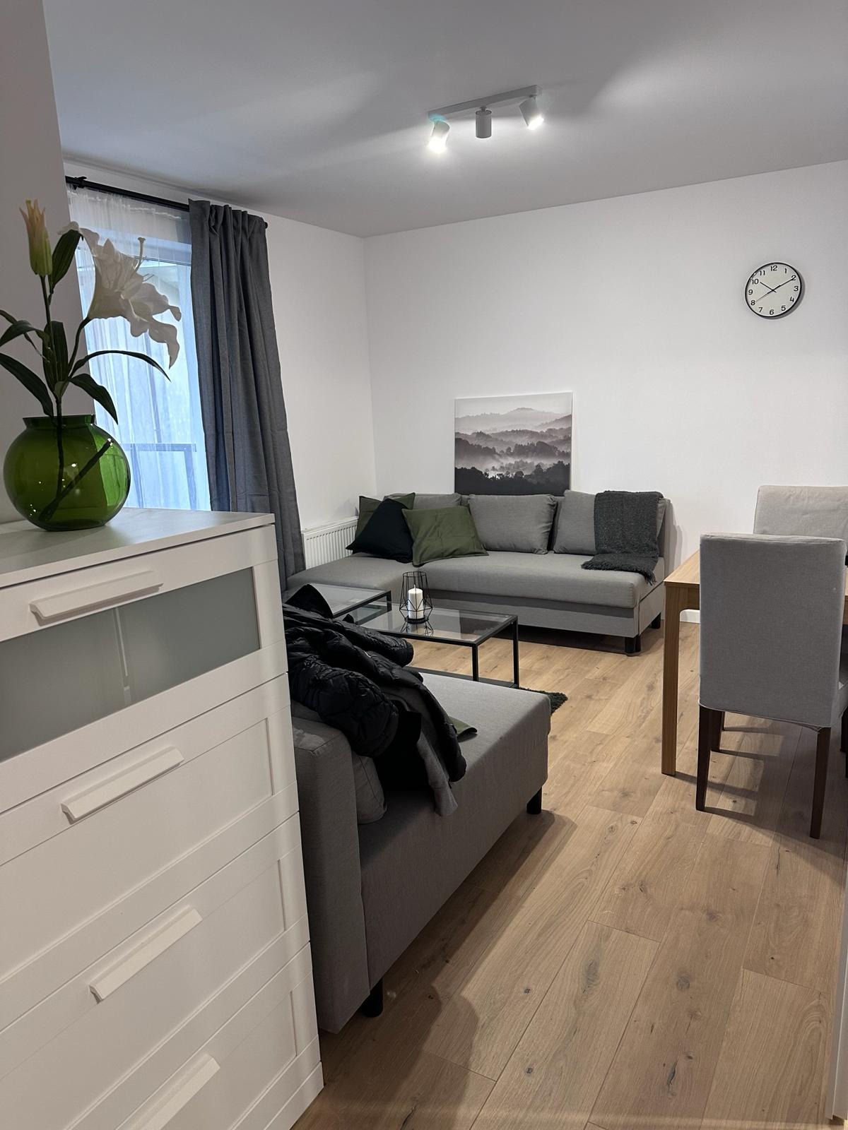 Jasne wnętrze salonu z drewnianą podłogą, szarą sofą, stolikiem kawowym i kwiatami w wazonie. Minimalistyczny wystrój z obrazem górskim i zegarem na ścianie.