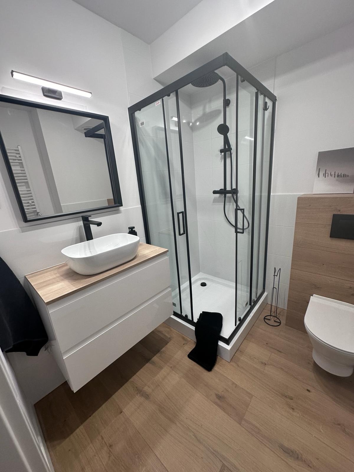 Nowoczesna łazienka z kabiną prysznicową w czarnej ramie, białą umywalką na szafce z drewnianym blatem i drewnopodobną okładziną ściany. Minimalistyczny design.